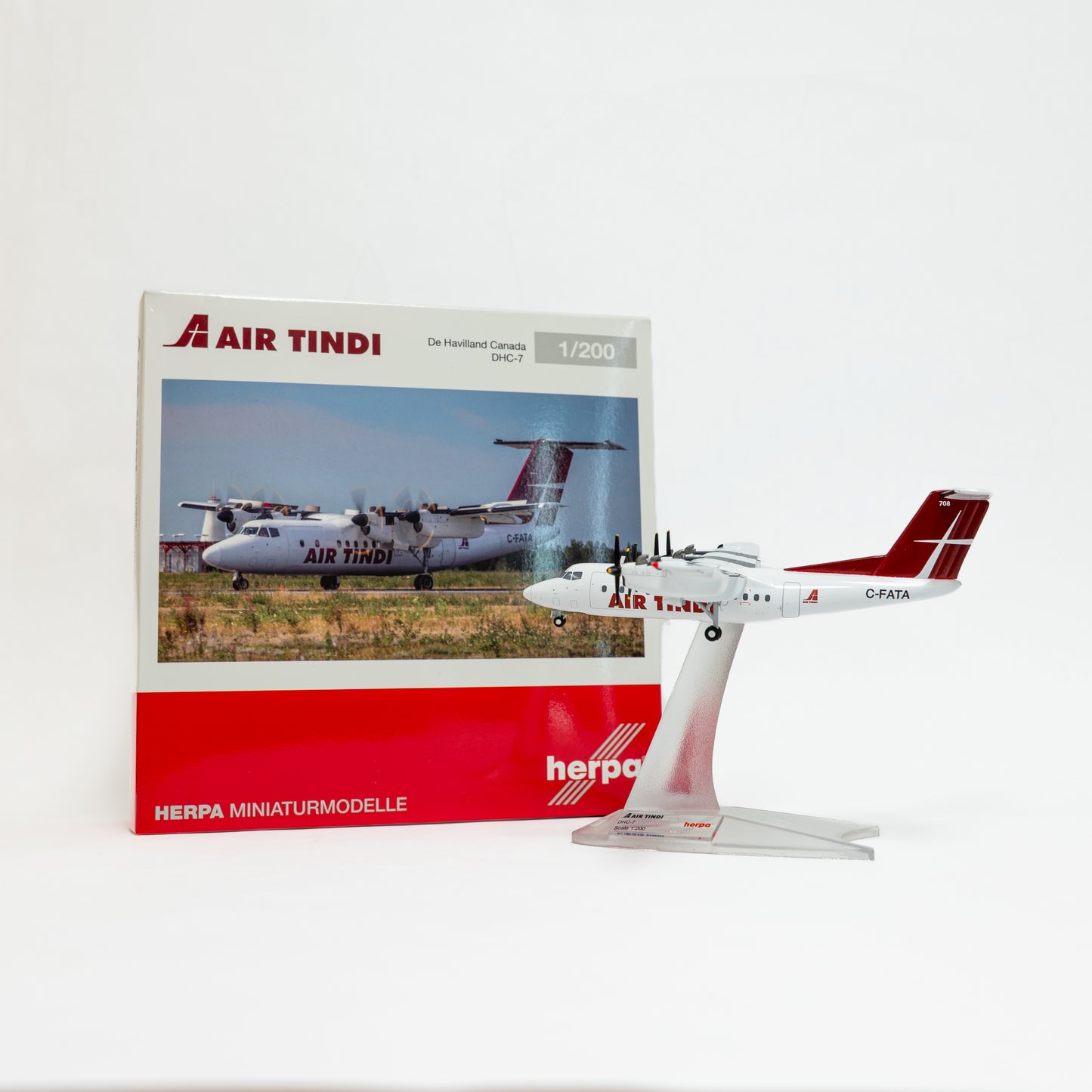 A 1:200 scale model airplane of an Air Tindi Dash 7 aircraft on a clear display stand, shown next to its original product box by Herpa Miniaturmodelle.