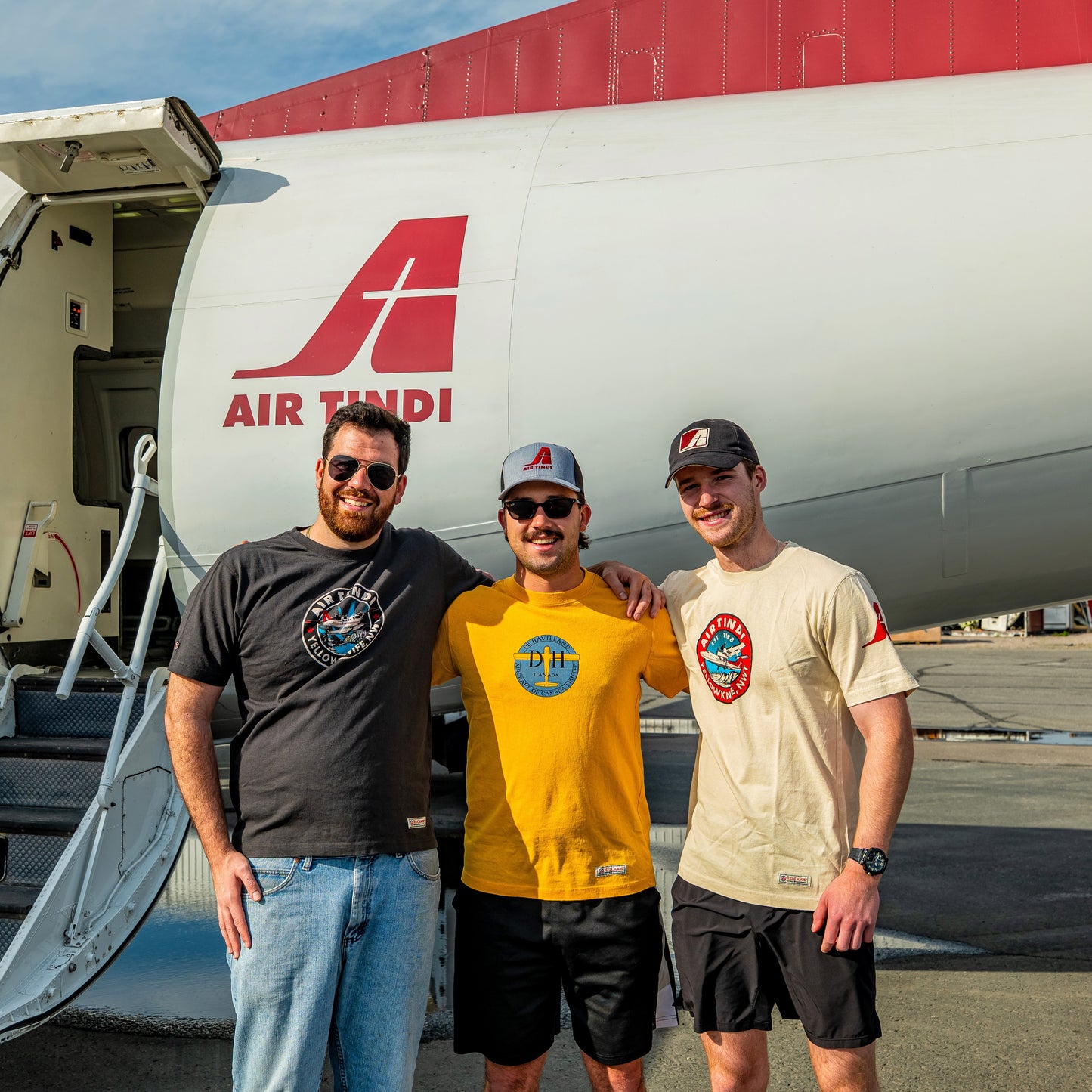 Model wearing the Limited Edition de Havilland Heritage Tee standing in front of an Air Tindi aircraft beside models wearing the Twin Otter Aurora Tee and Classic Dash 7 Tee