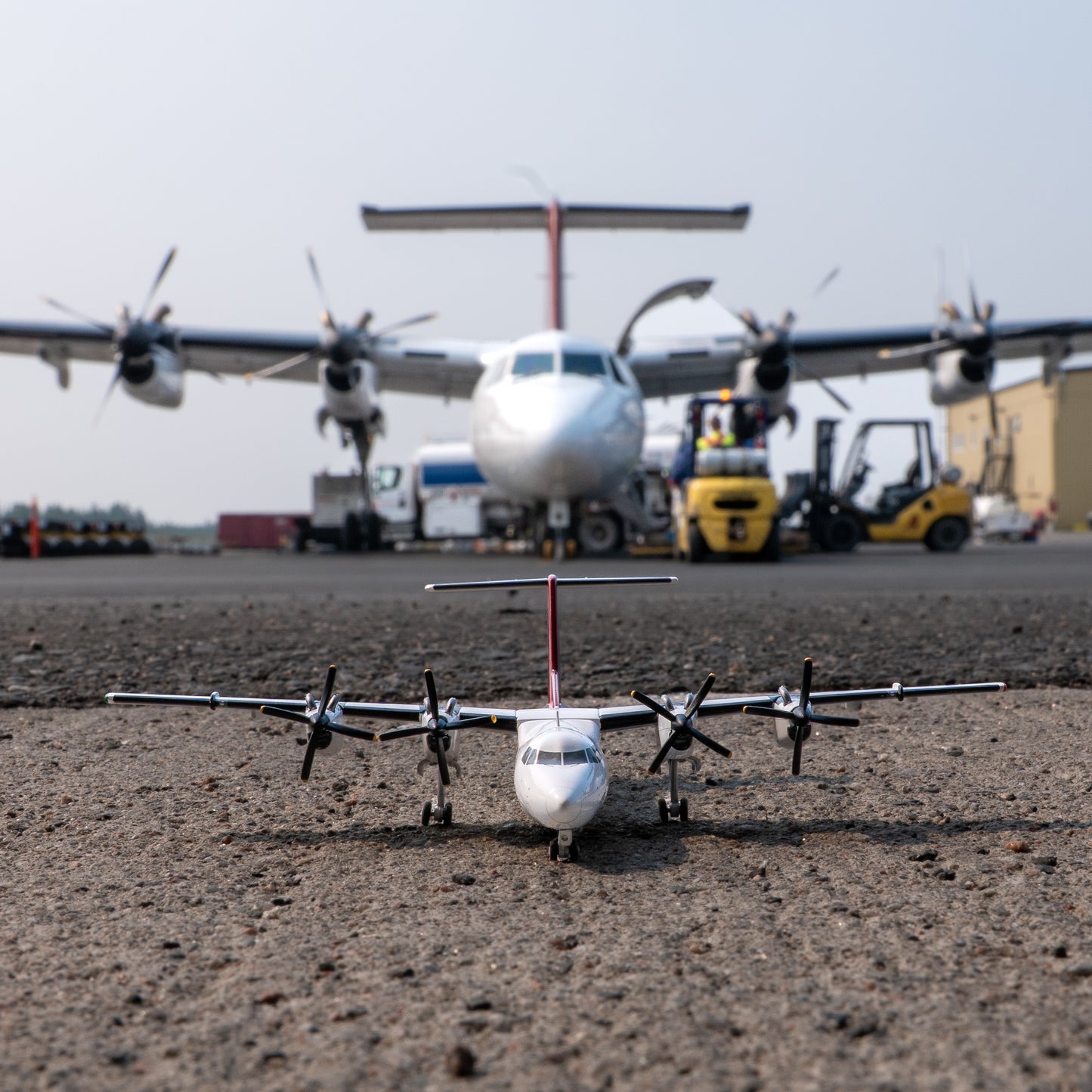 An Air Tindi Dash 7 model sits on tarmac in the foreground, with a larger real airplane and airport equipment blurred in the background on the tarmac.