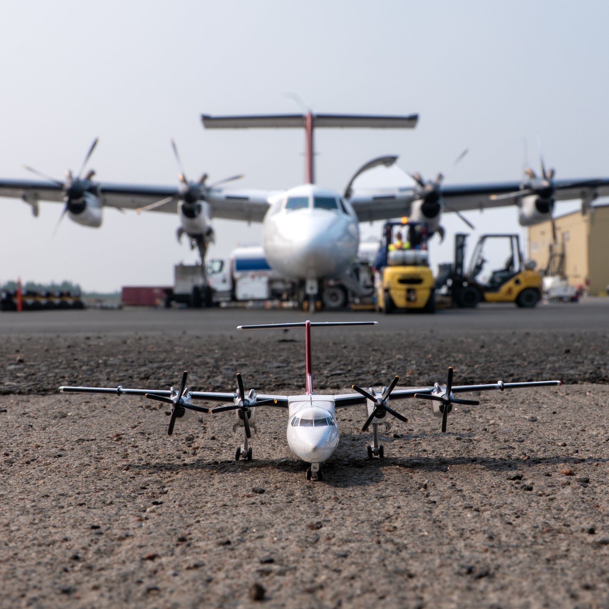 An Air Tindi Dash 7 model sits on tarmac in the foreground, with a larger real airplane and airport equipment blurred in the background on the tarmac.