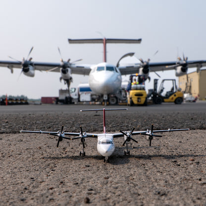 An Air Tindi Dash 7 model sits on tarmac in the foreground, with a larger real airplane and airport equipment blurred in the background on the tarmac.
