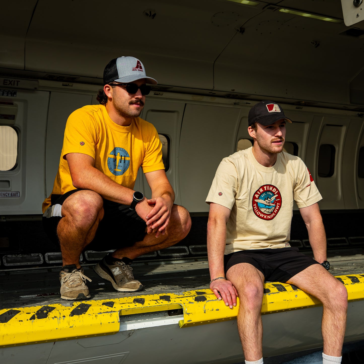 Model wearing the Limited Edition de Havilland Heritage Tee seated inside an aircraft beside a model wearing the Classic Dash 7 Tee
