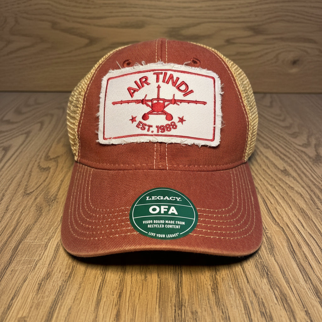 Air Tindi Legacy Trucker Hat in red and gold on a wooden tabletop background