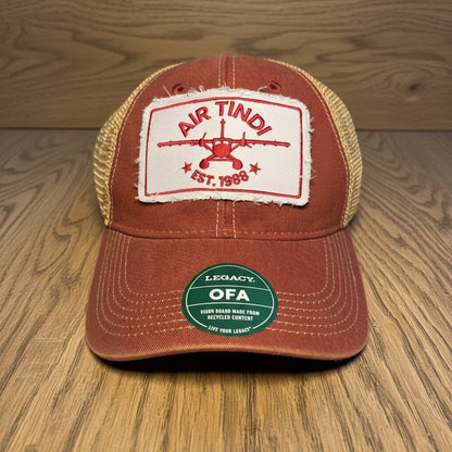 Air Tindi Legacy Trucker Hat in red and gold on a wooden tabletop background