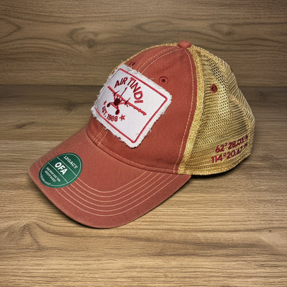 Air Tindi Legacy Trucker Hat in red and gold on a wooden tabletop background