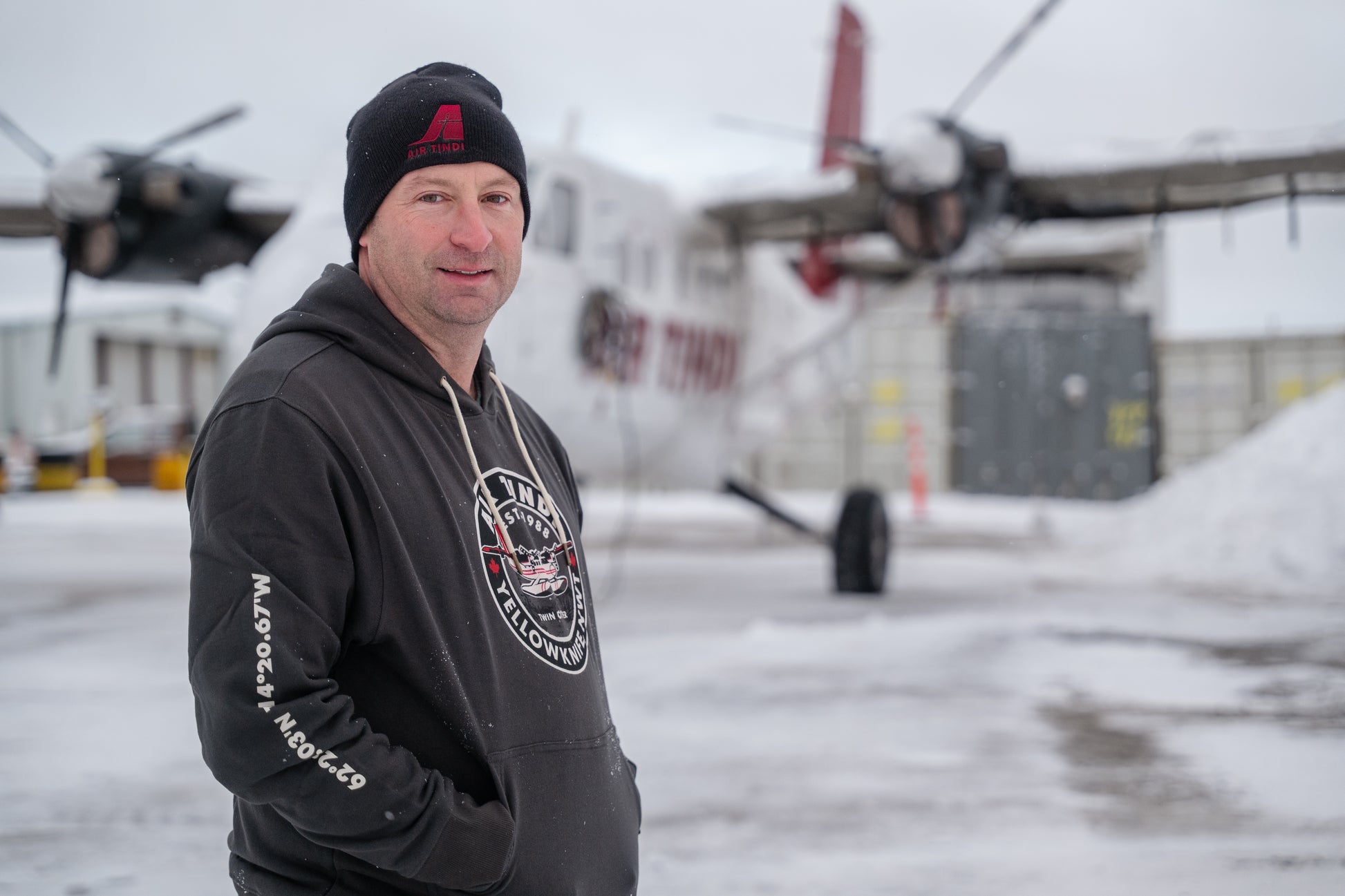 Model wearing an Air Tindi Twin Otter Hoodie in the snow, in front of a Twin Otter plane