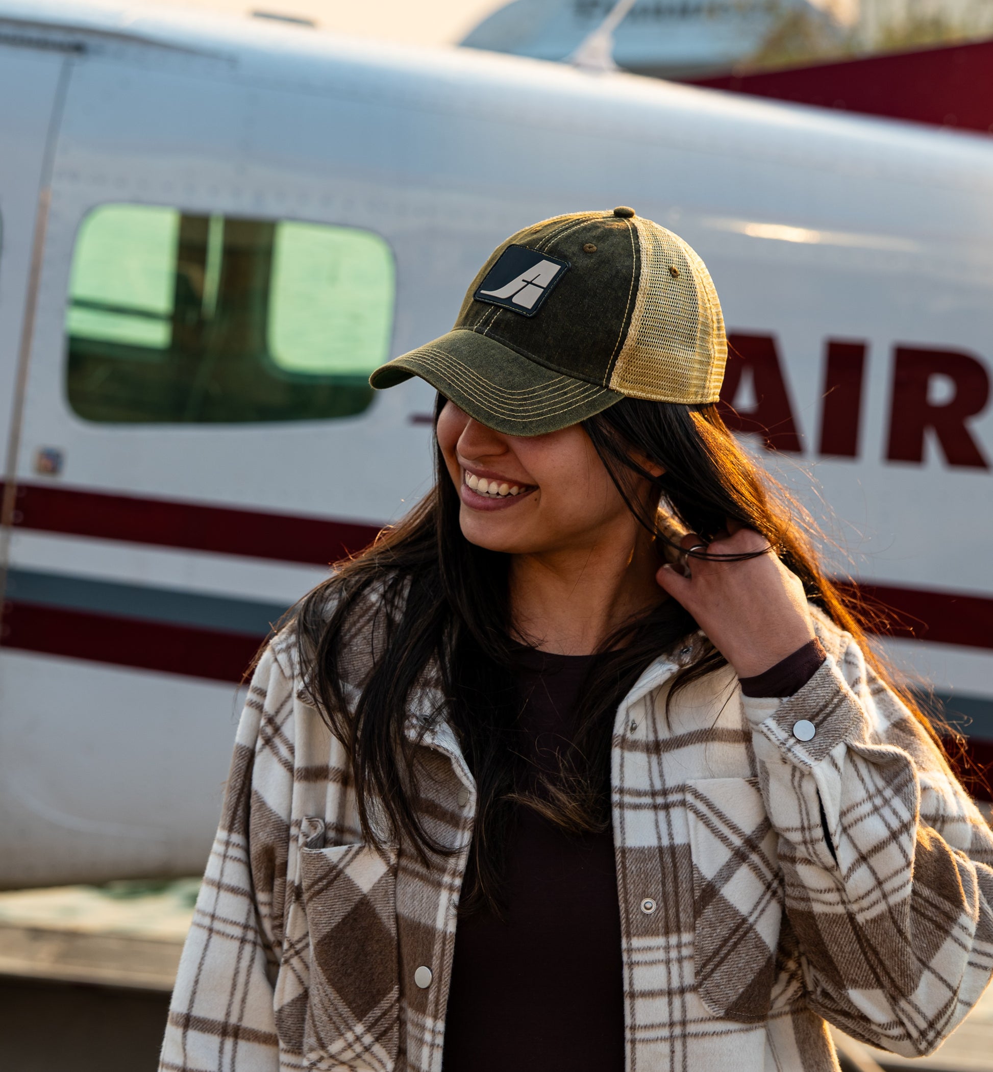 Model wearing a black and gold Air Tindi trucker hat in front of an aircraft