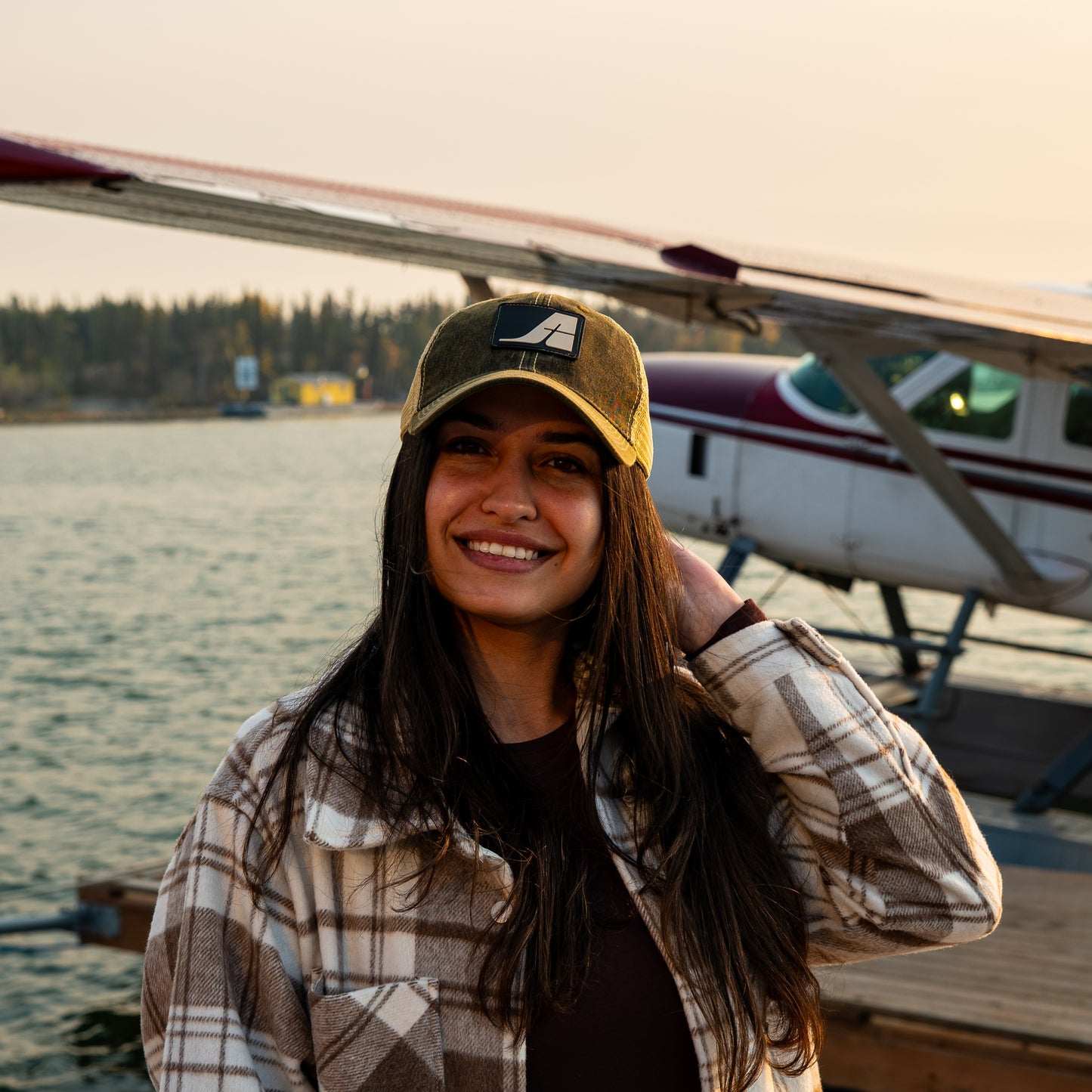 Model wearing a black and gold Air Tindi trucker hat in front of an aircraft and lake