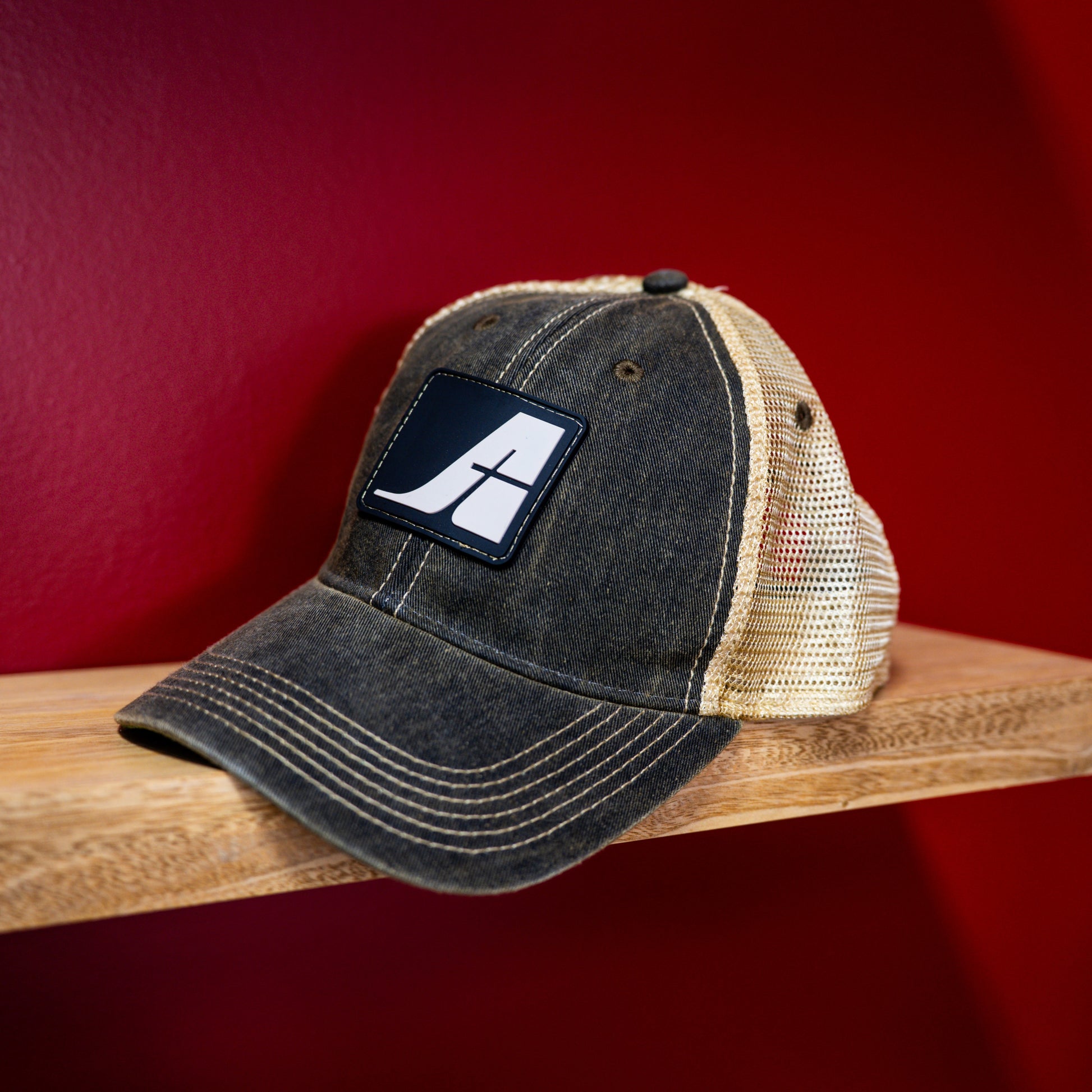 Black and gold Air Tindi trucker hat with a patch on a wooden surface against a red background